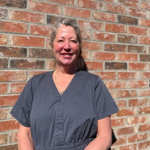 Woman with dark grey scrubs standing against brick wall, smiling, hair pulled back and arms crossed in front